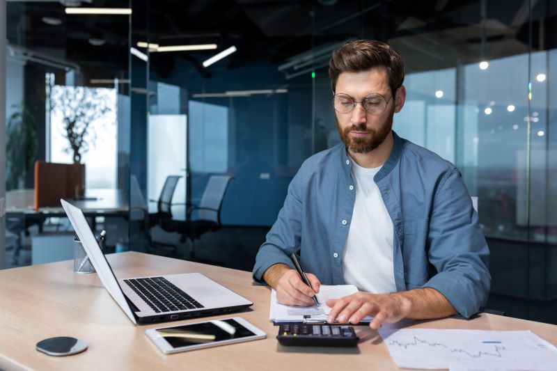 Homem utilizando calculadora com computador ao lado para realizar avaliações do ciclo de faturamento de sua empresa