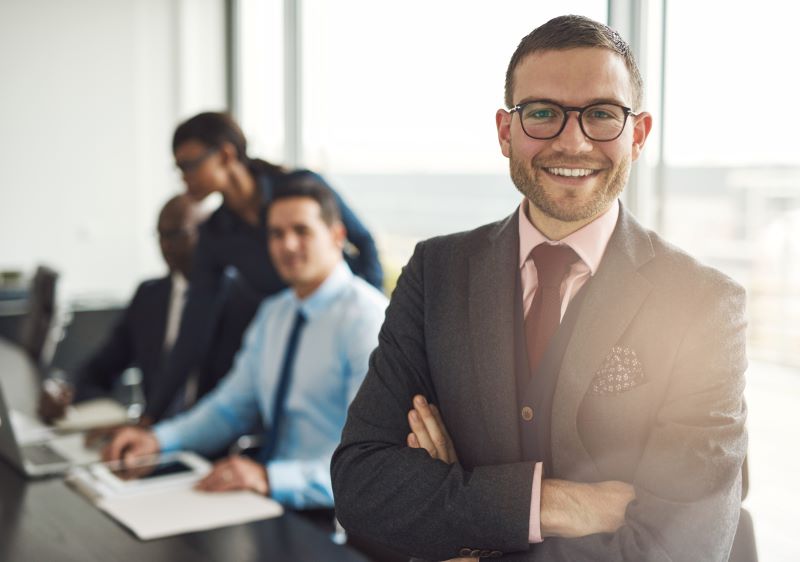 homem de social completo e óculos sorri enquanto olha para a câmera, com colegas de trabalho ao fundo em desfoque, ilustrando um cenário de gestão contábil