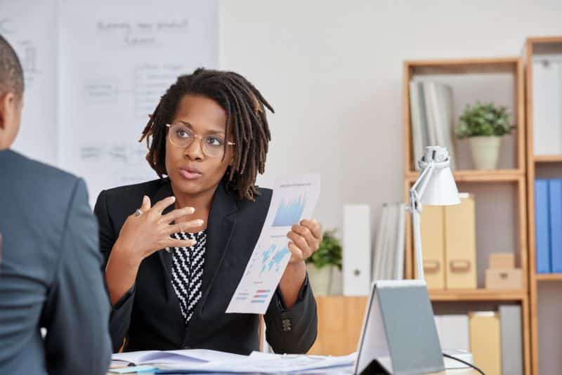 Mulher em escritório segurando relatório e explicando ao seu colega como a receita recorrente favorece a maximização de lucro.