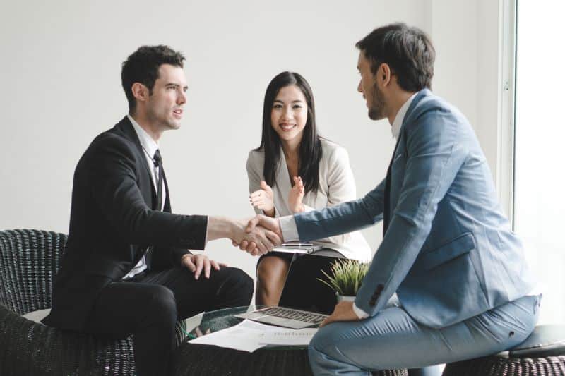 Vendedor em escritório apertando a mão de cliente enquanto colega ao lado aplaude, após um contrato fechado com cross sell e upsell.