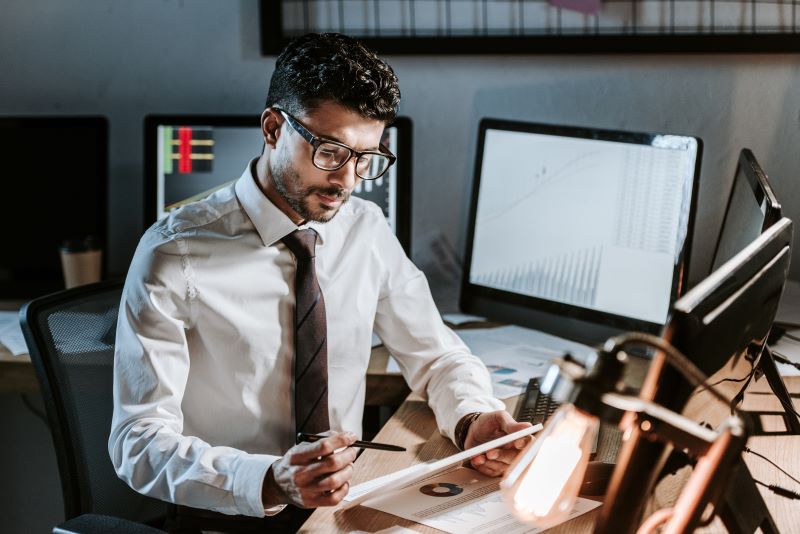 Homem em escritório com tablet e vários monitores analisando relatórios financeiros feitos com ajuda de tecnologias emergentes.