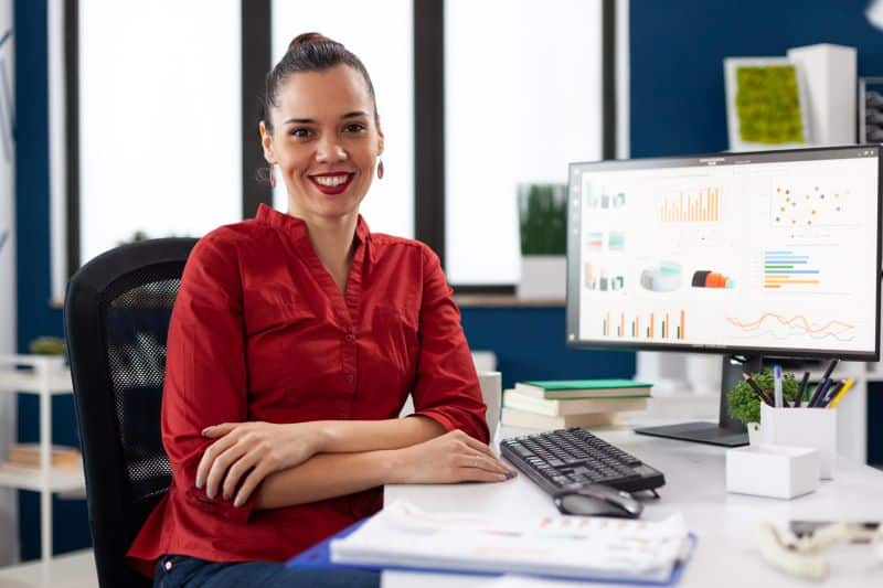 Mulher em escritório sorrindo para a câmera com sistema de gestão financeira aberto na tela do computador.